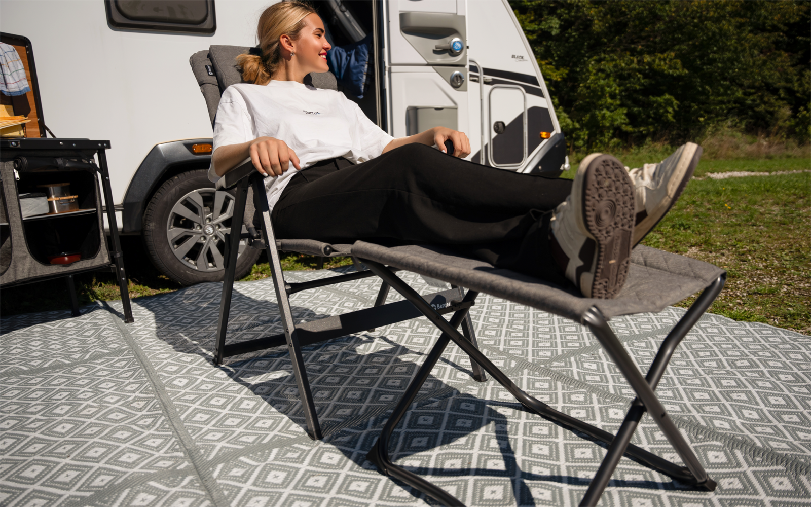 Person liegt auf einer zusammenklappbaren Campingliege vor einem Wohnmobil auf einem gemusterten Outdoor-Teppich, Bäume im Hintergrund