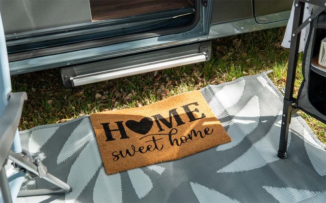 Paillasson brun en coco avec inscription noire "HOME sweet home" et symbole de cœur, posé sur un tapis d'extérieur gris-blanc devant une porte de véhicule ouverte ; herbe, marche métallique et pied de chaise visibles