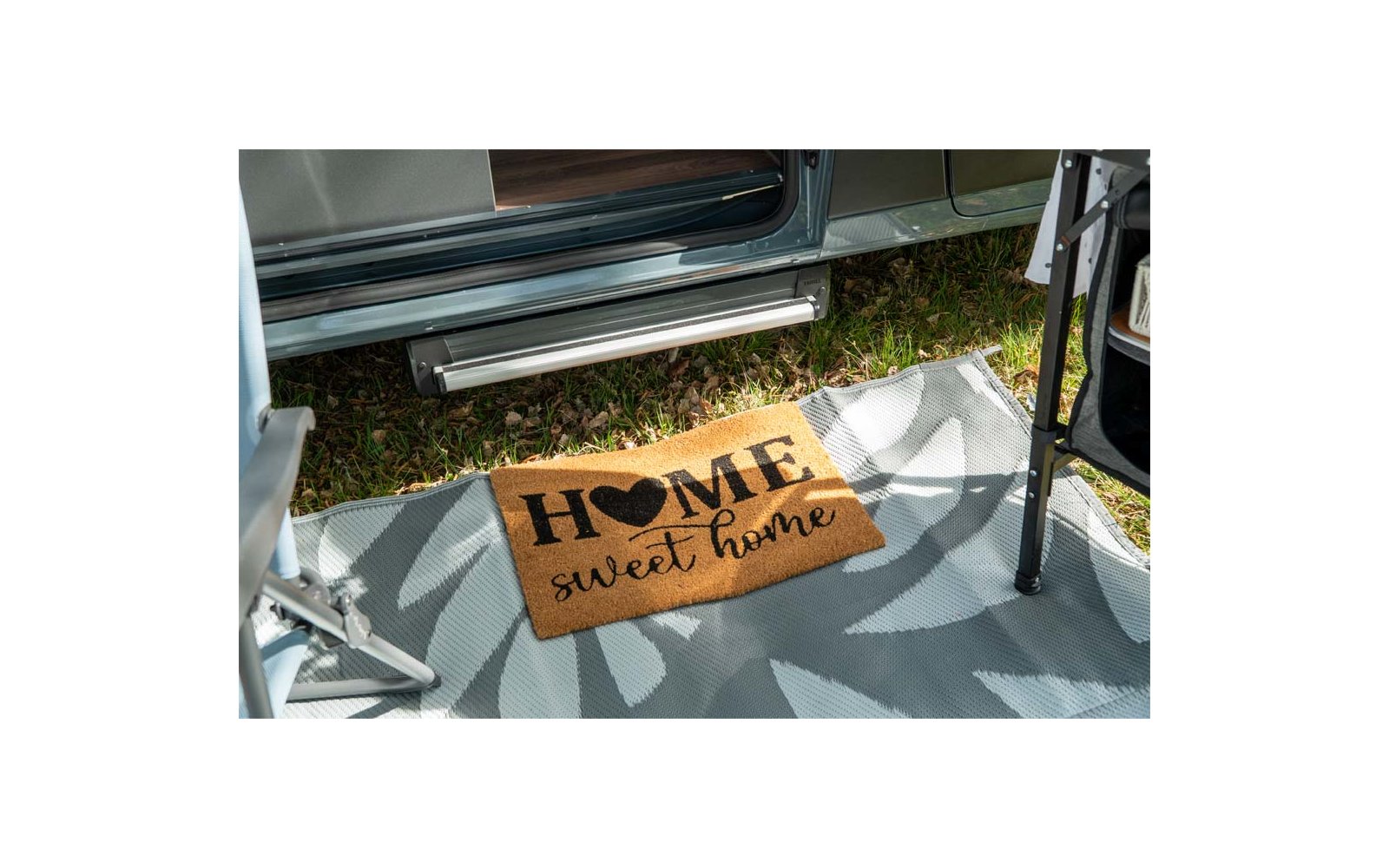Paillasson brun en coco avec inscription noire "HOME sweet home" et symbole de cœur, posé sur un tapis d'extérieur gris-blanc devant une porte de véhicule ouverte ; herbe, marche métallique et pied de chaise visibles
