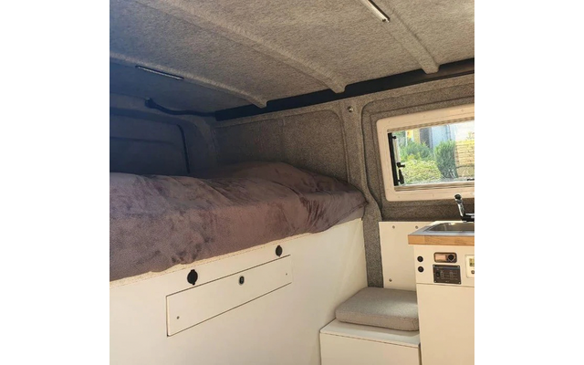 Voertuiginterieur: gewatteerd ligbed met deken, wit opbergmeubel met lade en ronde openingen, klein raam met planten buiten, werkblad met gootsteen en kraan, gewatteerde zitplaats, grijze stoffen wand- en plafondpanelen