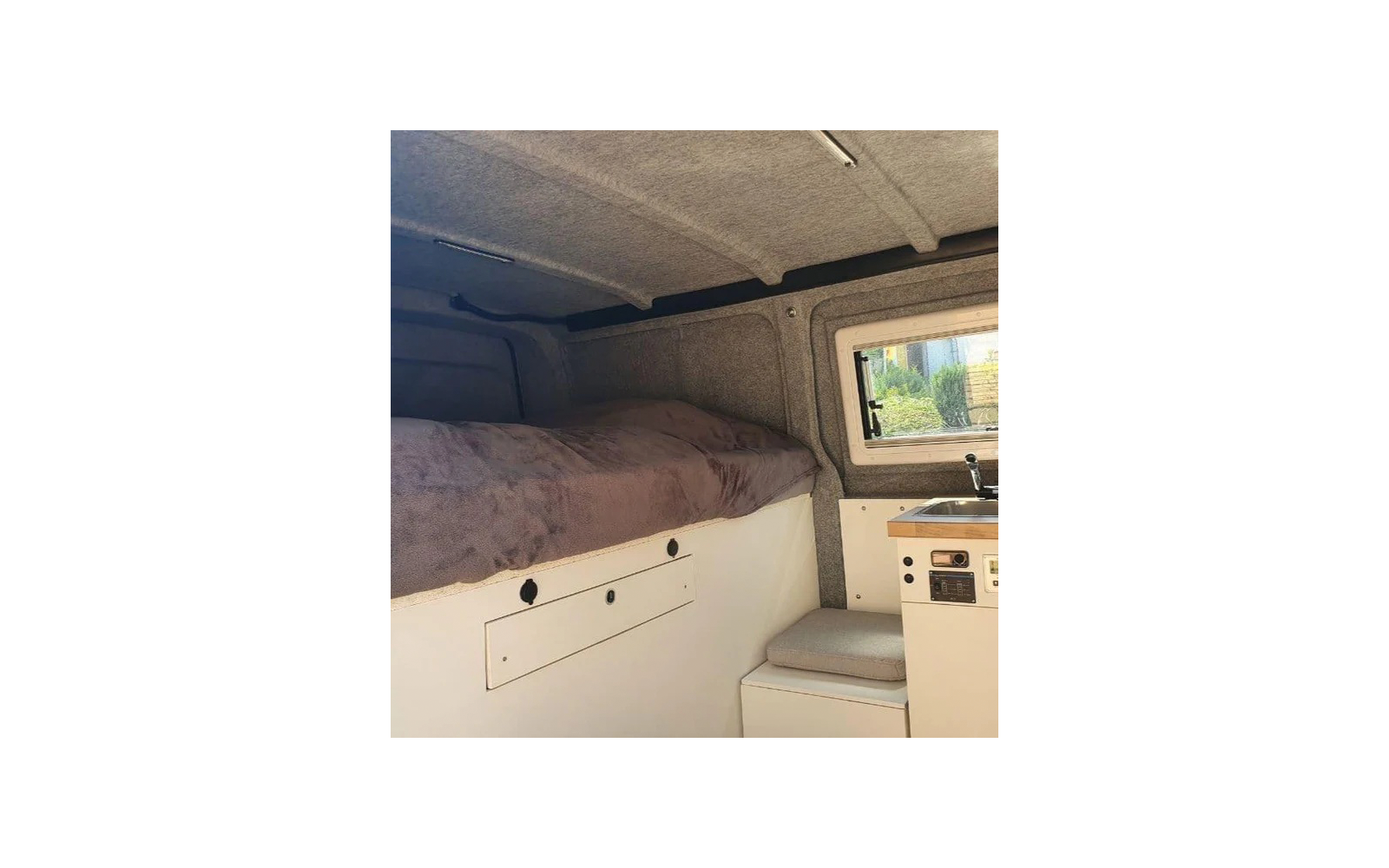 Voertuiginterieur: gewatteerd ligbed met deken, wit opbergmeubel met lade en ronde openingen, klein raam met planten buiten, werkblad met gootsteen en kraan, gewatteerde zitplaats, grijze stoffen wand- en plafondpanelen