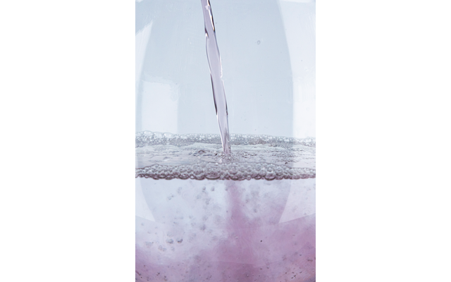 Nahaufnahme eines Flüssigkeitsstrahls in ein Glas mit hellvioletter Flüssigkeit und schäumenden Blasen auf der Oberfläche