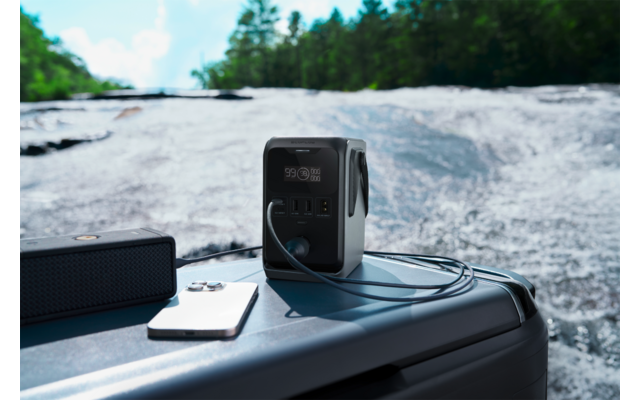 Draagbare stroomgenerator op een grijze kist aan de rand van een stromende rivier; daarnaast een witte smartphone, een zwarte draagbare speaker en een kabel