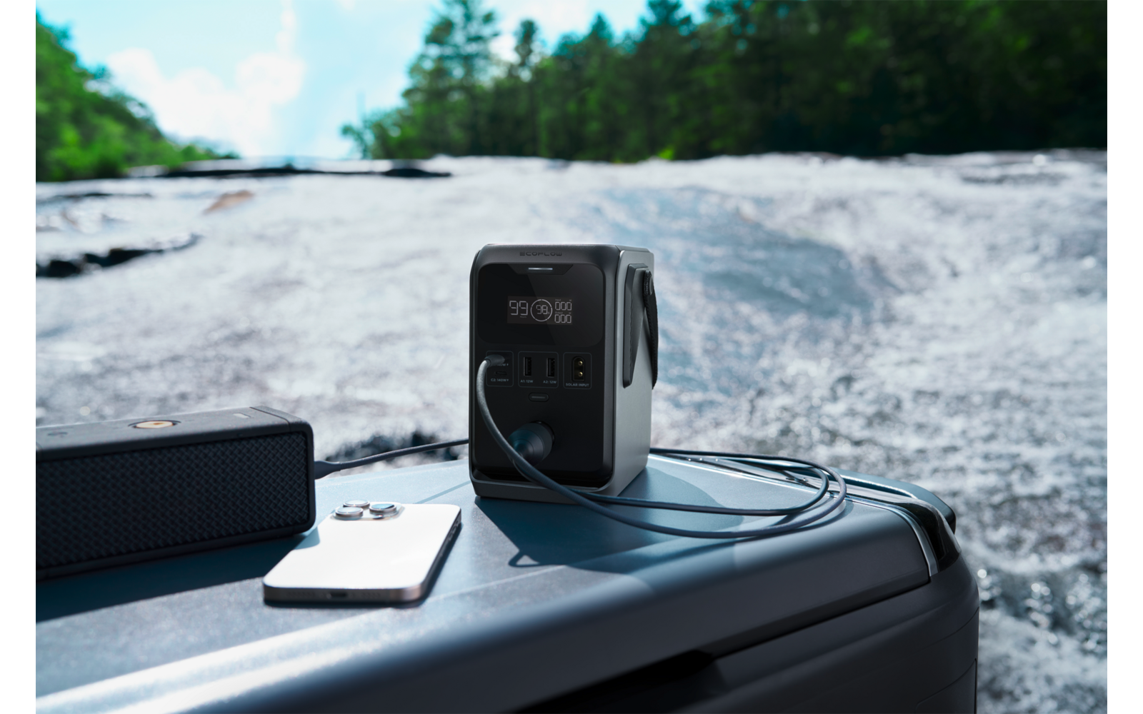 Draagbare stroomgenerator op een grijze kist aan de rand van een stromende rivier; daarnaast een witte smartphone, een zwarte draagbare speaker en een kabel
