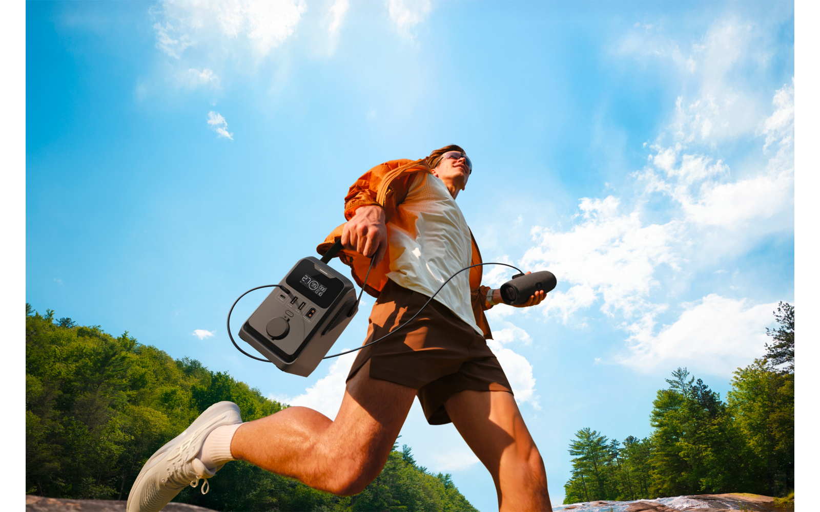 Persoon springt over rotsen, draagt oranje rugzak en witte sportschoenen, houdt een grijs apparaat met kabel en oortelefoon; blauwe lucht en bomen op de achtergrond