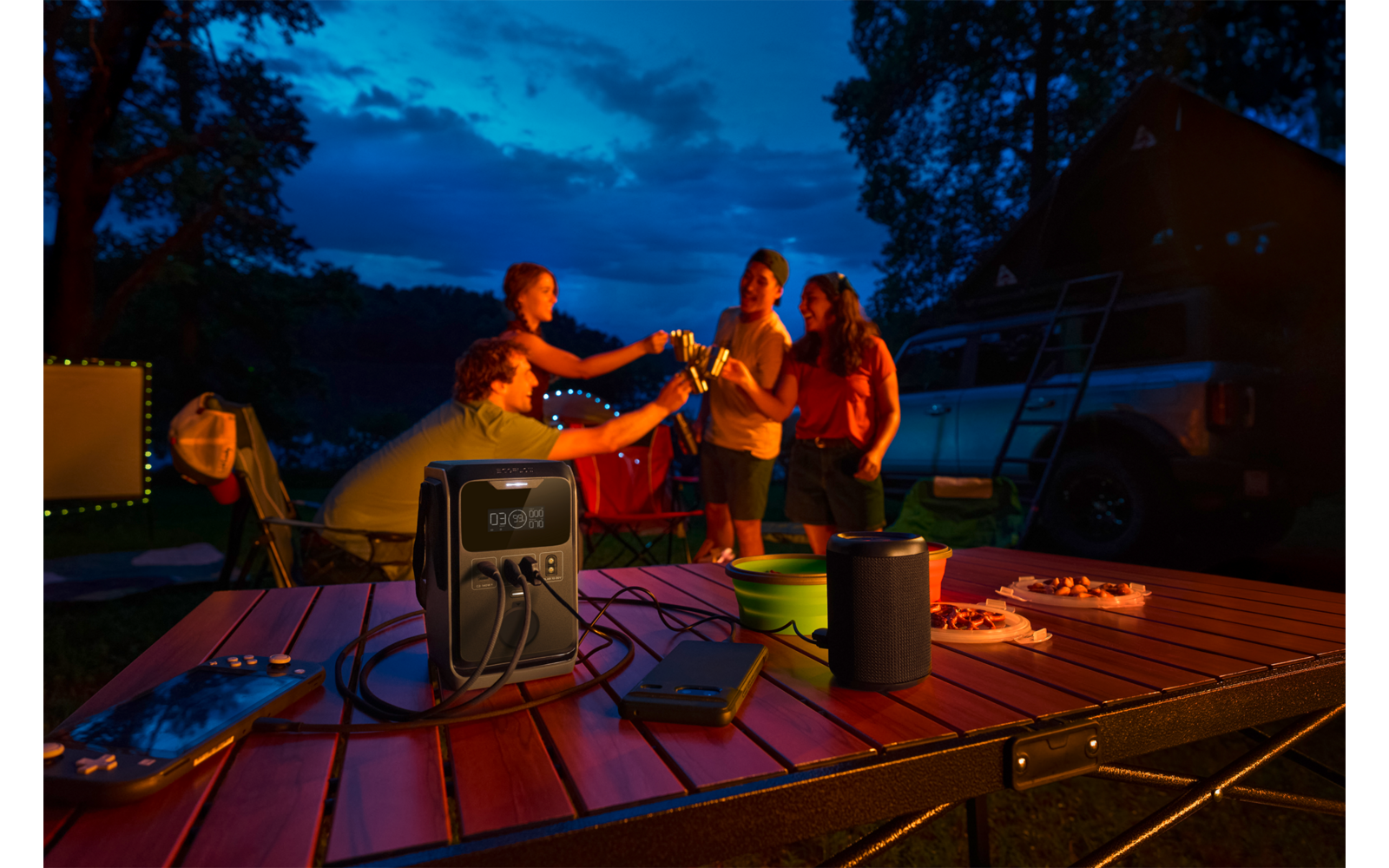 Vijf personen bij een kampeertafel in de schemering, proostend met drankjes; op de tafel een draagbare stroomvoorziening met kabels, een luidspreker, borden met hapjes en een smartphone; op de achtergrond een camper, bomen en een donkere lucht