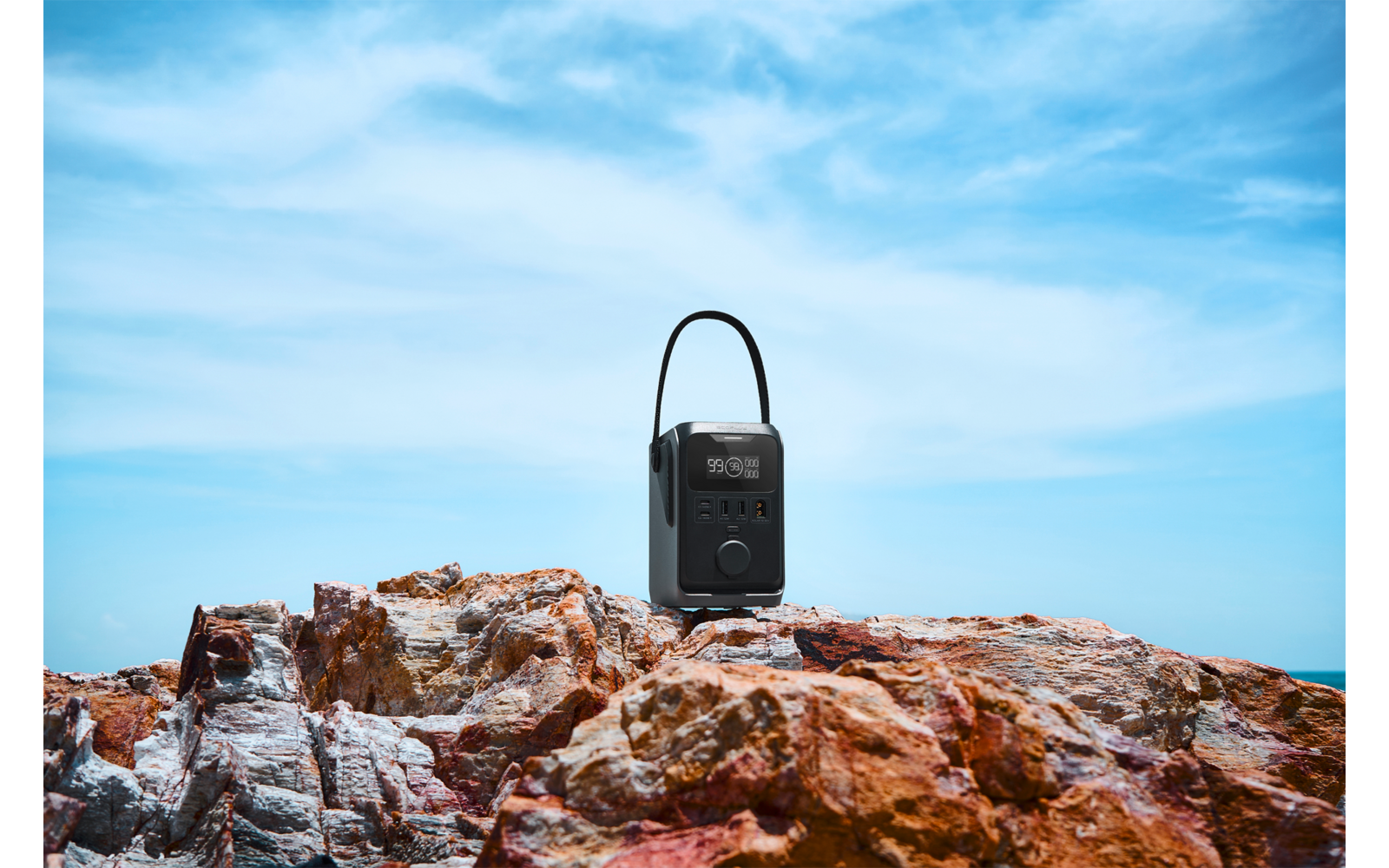 Zwart draagbaar apparaat met handvat op roodbruine rotsen, op de achtergrond zee en blauwe lucht met wolken