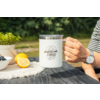 Main avec montre tient une tasse blanche avec le logo Camp Life sur une table en bois, à côté une moitié de citron, un citron entier, un petit bol de baies et une plante en pot, arrière-plan de jardin flou