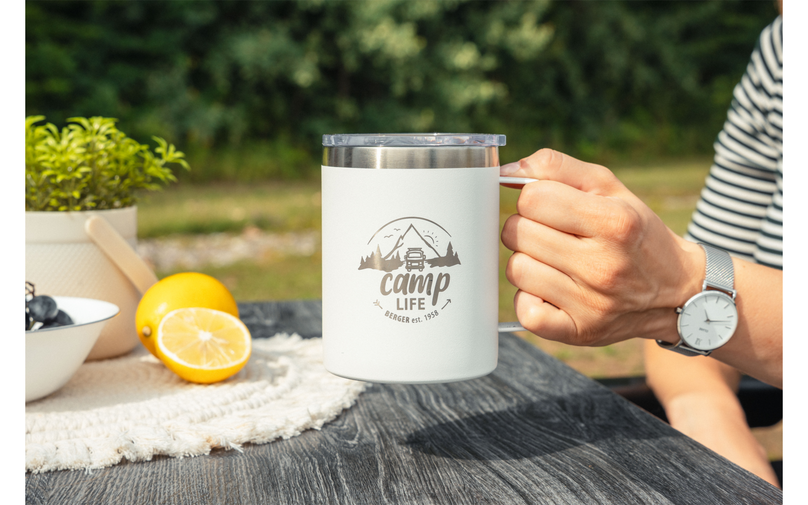 Main avec montre tient une tasse blanche avec le logo Camp Life sur une table en bois, à côté une moitié de citron, un citron entier, un petit bol de baies et une plante en pot, arrière-plan de jardin flou