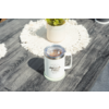 Tasse blanche avec anse et couvercle transparent portant l'inscription "camp life" sur une surface en bois grise ; en arrière-plan un rond de table crocheté et un petit pot avec plante verte