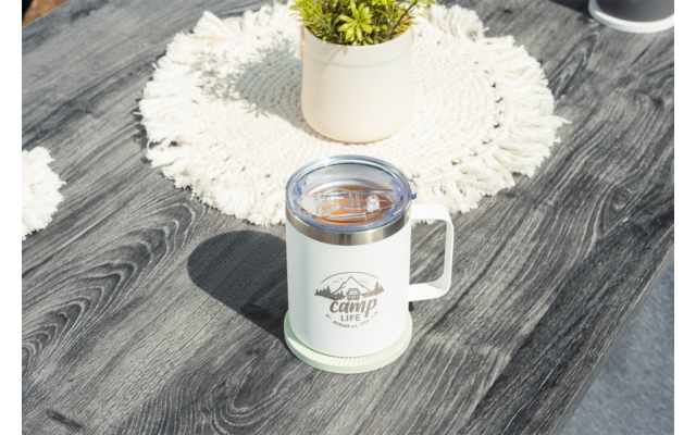 Tasse blanche avec anse et couvercle transparent portant l'inscription "camp life" sur une surface en bois grise ; en arrière-plan un rond de table crocheté et un petit pot avec plante verte