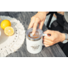 Deux mains tiennent un couvercle transparent au-dessus d'une tasse isotherme blanche avec le logo « camp life » sur une table en bois foncé ; un demi-citron et un citron entier sont posés sur un napperon rond tressé ; une bague et une montre sont visibles sur les mains