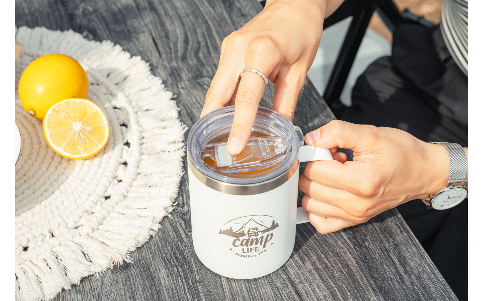 Deux mains tiennent un couvercle transparent au-dessus d'une tasse isotherme blanche avec le logo « camp life » sur une table en bois foncé ; un demi-citron et un citron entier sont posés sur un napperon rond tressé ; une bague et une montre sont visibles sur les mains