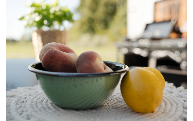 Bol vert contenant plusieurs pêches sur une nappe claire et texturée ; un citron jaune à droite ; pot de plante et barbecue flous en arrière-plan