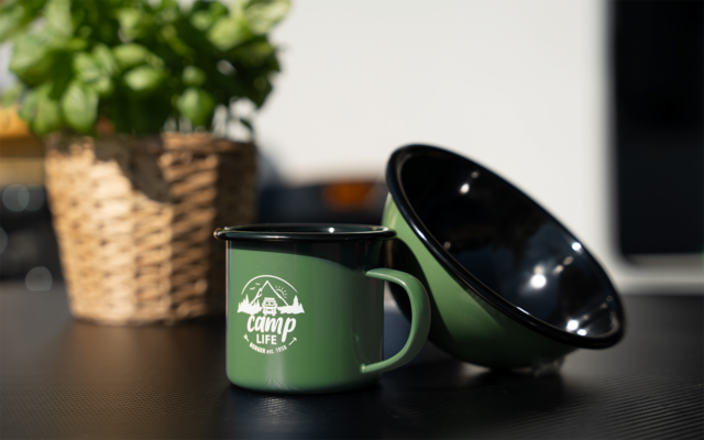 Tasse émaillée verte avec inscription blanche 'camp life' à côté d'un bol émaillé vert incliné; en arrière-plan un panier tressé avec une plante sur une table noire