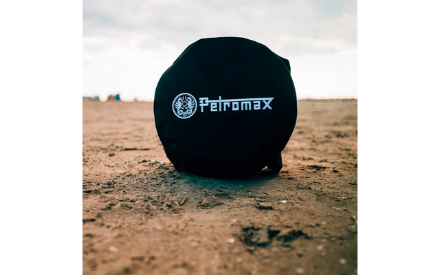 Rundes schwarzes Etui mit weißem 'Petromax'-Schriftzug und Logo auf sandigem Boden, unscharfer Horizont und bewölkter Himmel