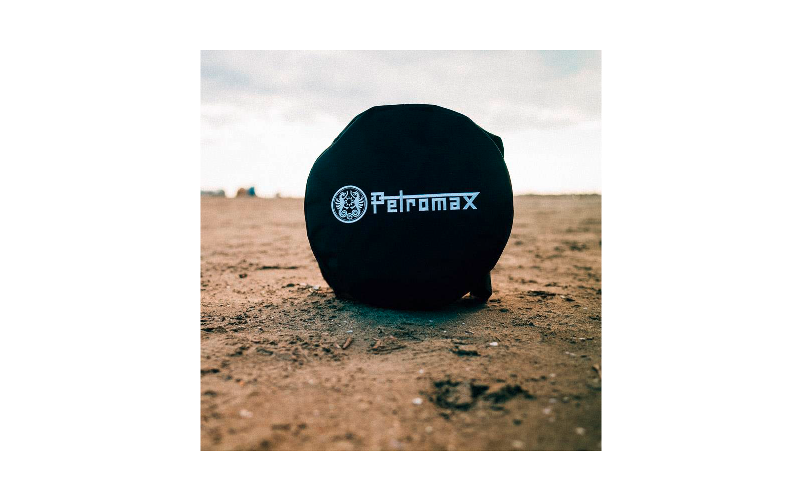 Rundes schwarzes Etui mit weißem 'Petromax'-Schriftzug und Logo auf sandigem Boden, unscharfer Horizont und bewölkter Himmel