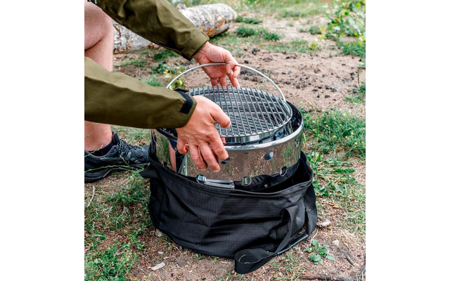 Hände einer Person setzen einen runden Edelstahlgrill mit Grillrost in eine schwarze Transporttasche auf Gras
