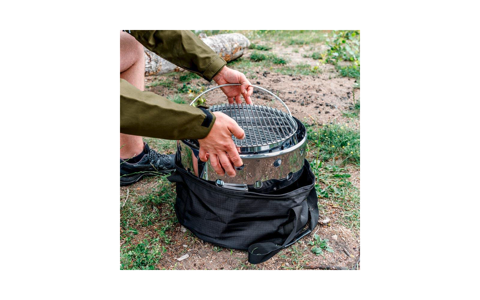 Hände einer Person setzen einen runden Edelstahlgrill mit Grillrost in eine schwarze Transporttasche auf Gras