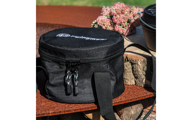 Schwarze, runde Tasche mit Reißverschluss, Tragegriffen und weißem Aufdruck, auf einer Holzfläche vor gestapeltem Holz; im Hintergrund ein gusseiserner Topf und rosa Blüten