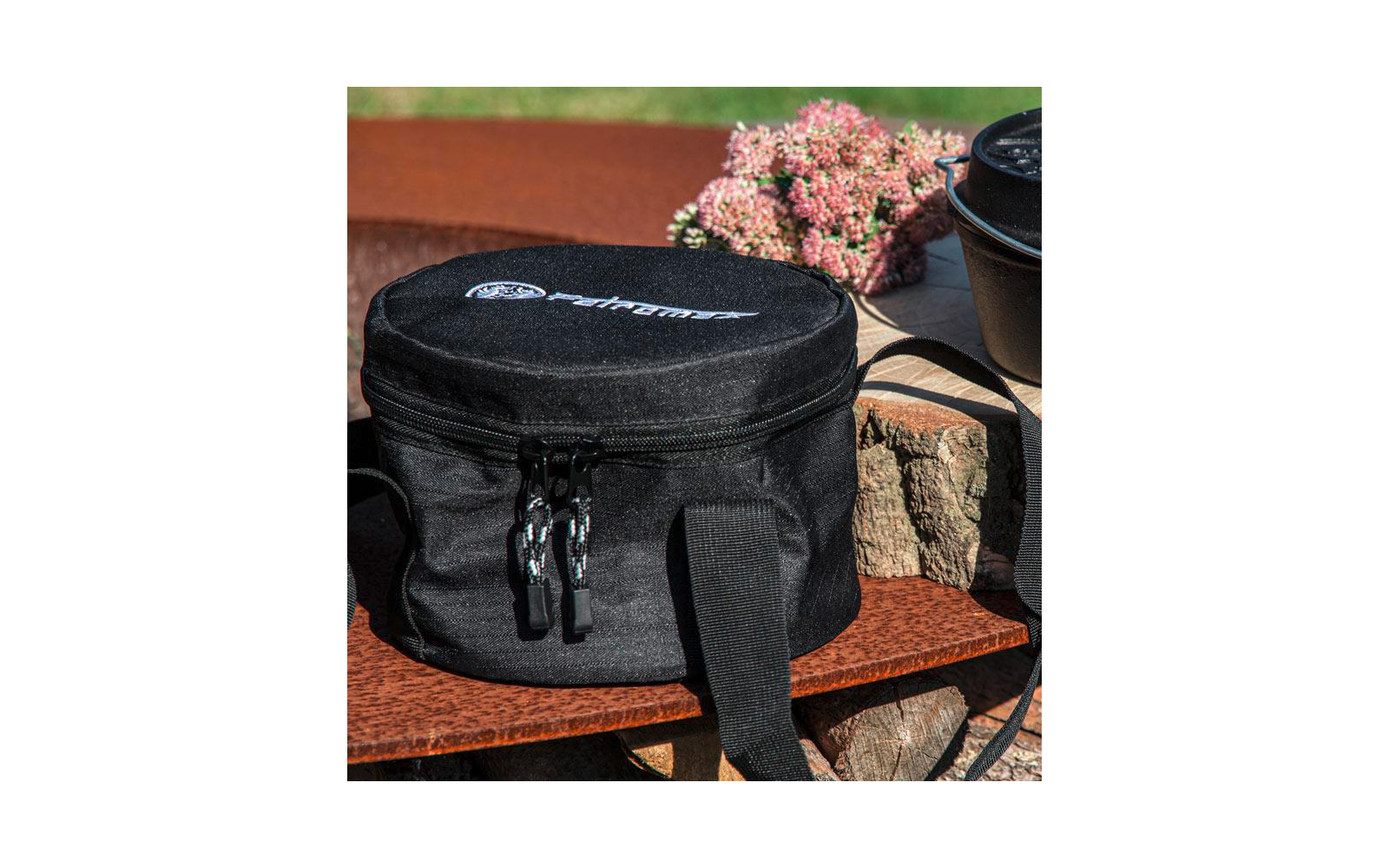 Schwarze, runde Tasche mit Reißverschluss, Tragegriffen und weißem Aufdruck, auf einer Holzfläche vor gestapeltem Holz; im Hintergrund ein gusseiserner Topf und rosa Blüten