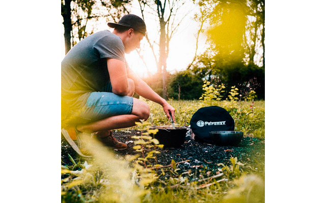 Person mit rückwärts getragenem Baseballcap hockt auf einer Wiese und rührt mit einem Löffel in einem schwarzen Topf; daneben ein weiterer Topf und eine schwarze runde Tasche mit weißem Logo
