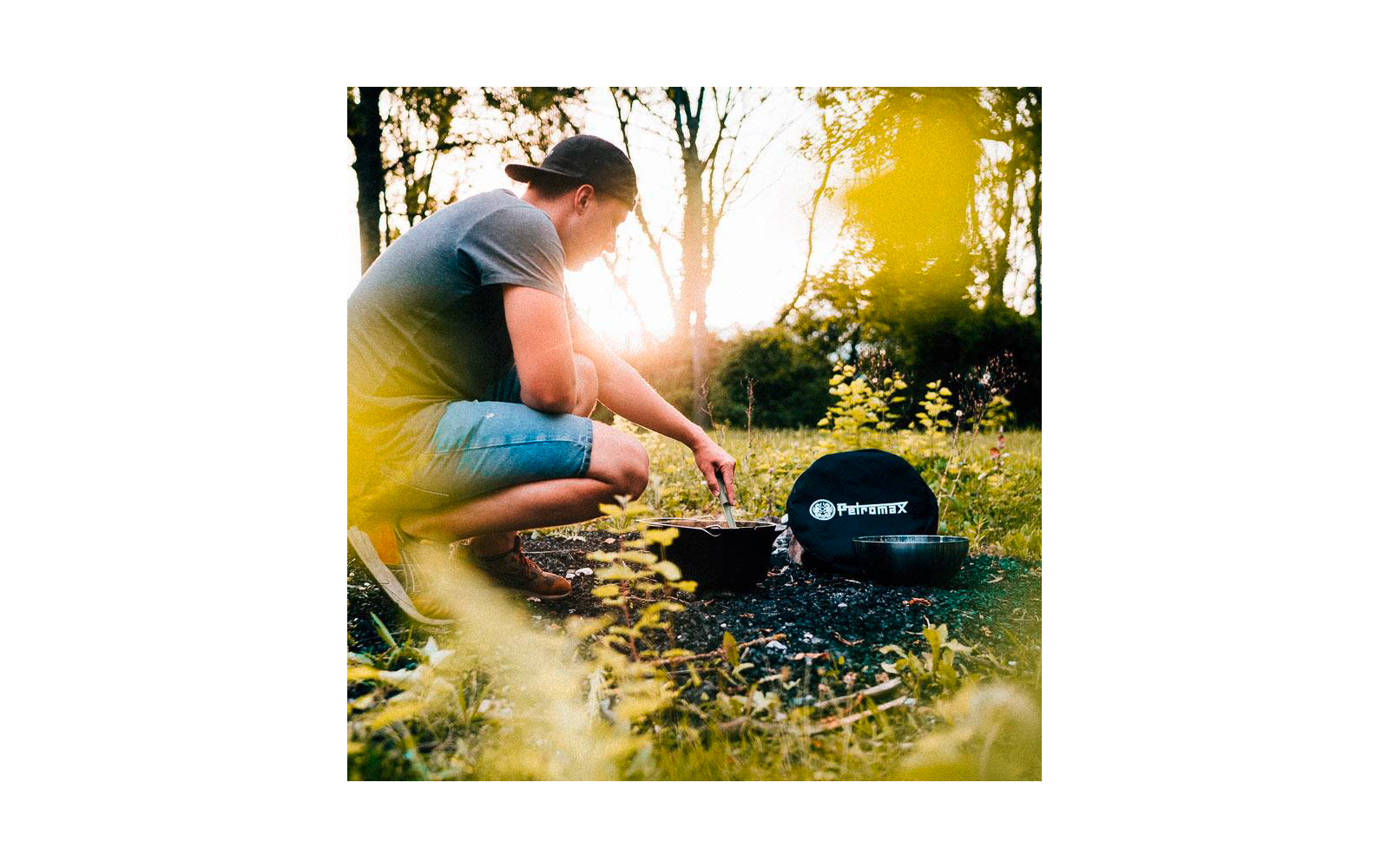 Person mit rückwärts getragenem Baseballcap hockt auf einer Wiese und rührt mit einem Löffel in einem schwarzen Topf; daneben ein weiterer Topf und eine schwarze runde Tasche mit weißem Logo
