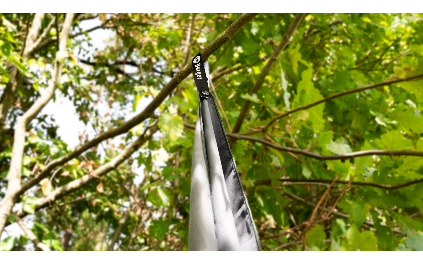 Zusammengeklappter schwarzer Regenschirm, an seiner Trageschlaufe an einem dünnen Ast aufgehängt, grüne Blätter im Hintergrund