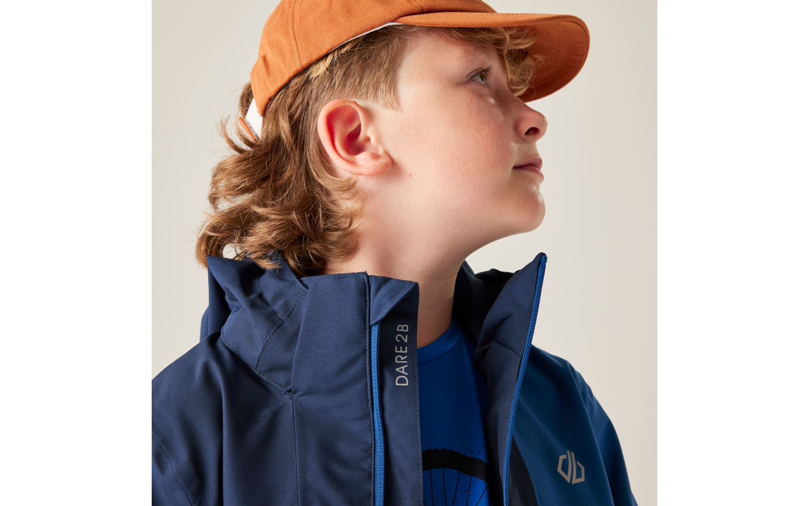 Person im Profil mit orangefarbener Kappe, welligen hellbraunen Haaren, dunkelblauer Jacke mit Reißverschluss und Schriftzug 'DARE2B', blaues Shirt mit rundem dunklem Aufdruck vor hellem Hintergrund