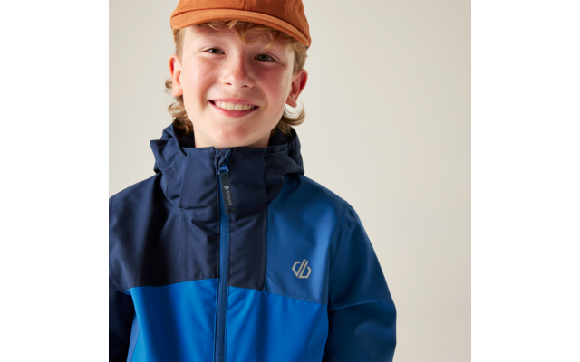 Nahaufnahme einer lächelnden Person mit orangefarbener Kappe und zweifarbiger blauer Jacke mit Reißverschluss und Brustlogo vor hellem Hintergrund