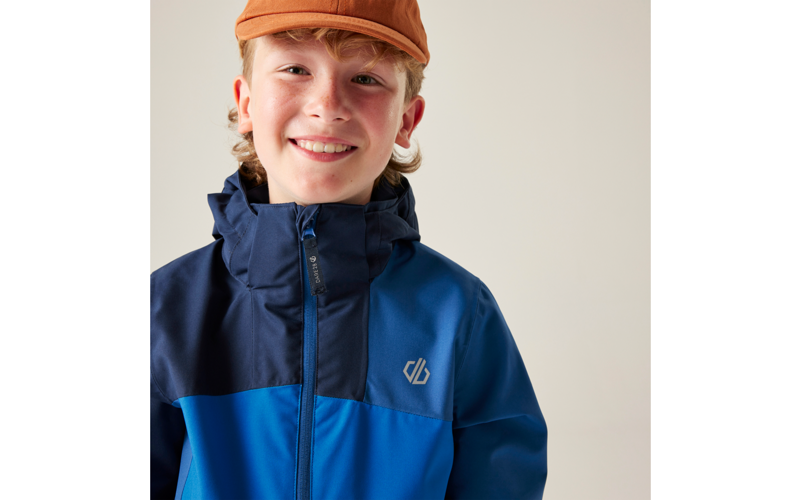 Nahaufnahme einer lächelnden Person mit orangefarbener Kappe und zweifarbiger blauer Jacke mit Reißverschluss und Brustlogo vor hellem Hintergrund