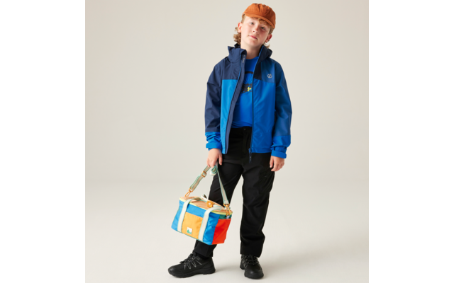 Person mit orangefarbener Mütze, zweifarbiger blauer Jacke, schwarzer Hose und schwarzen Schuhen steht vor hellem Hintergrund und hält eine bunte Tasche in einer Hand