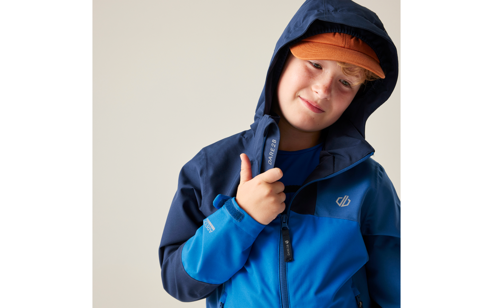 Person mit blauer, zweifarbiger Kapuzenjacke und orangefarbener Kappe, greift am Reißverschluss, heller Hintergrund