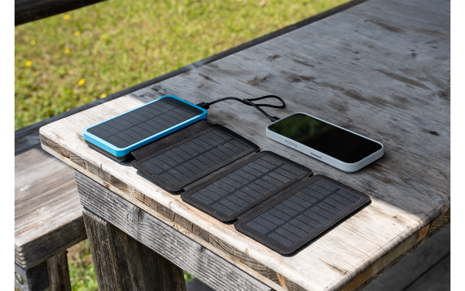 Ausgeklapptes tragbares Solarpanel auf einem Holztisch neben zwei mobilen Geräten, durch ein Kabel verbunden; im Hintergrund Rasen