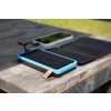 Rechteckiges Gerät mit schwarzer Solarfläche, blauem Rahmen und Lederlasche; dahinter gefaltetes schwarzes Solarpanel und weißes Smartphone mit schwarzem Bildschirm; alles auf einem verwitterten Holztisch; unscharfer grüner Rasen im Hintergrund