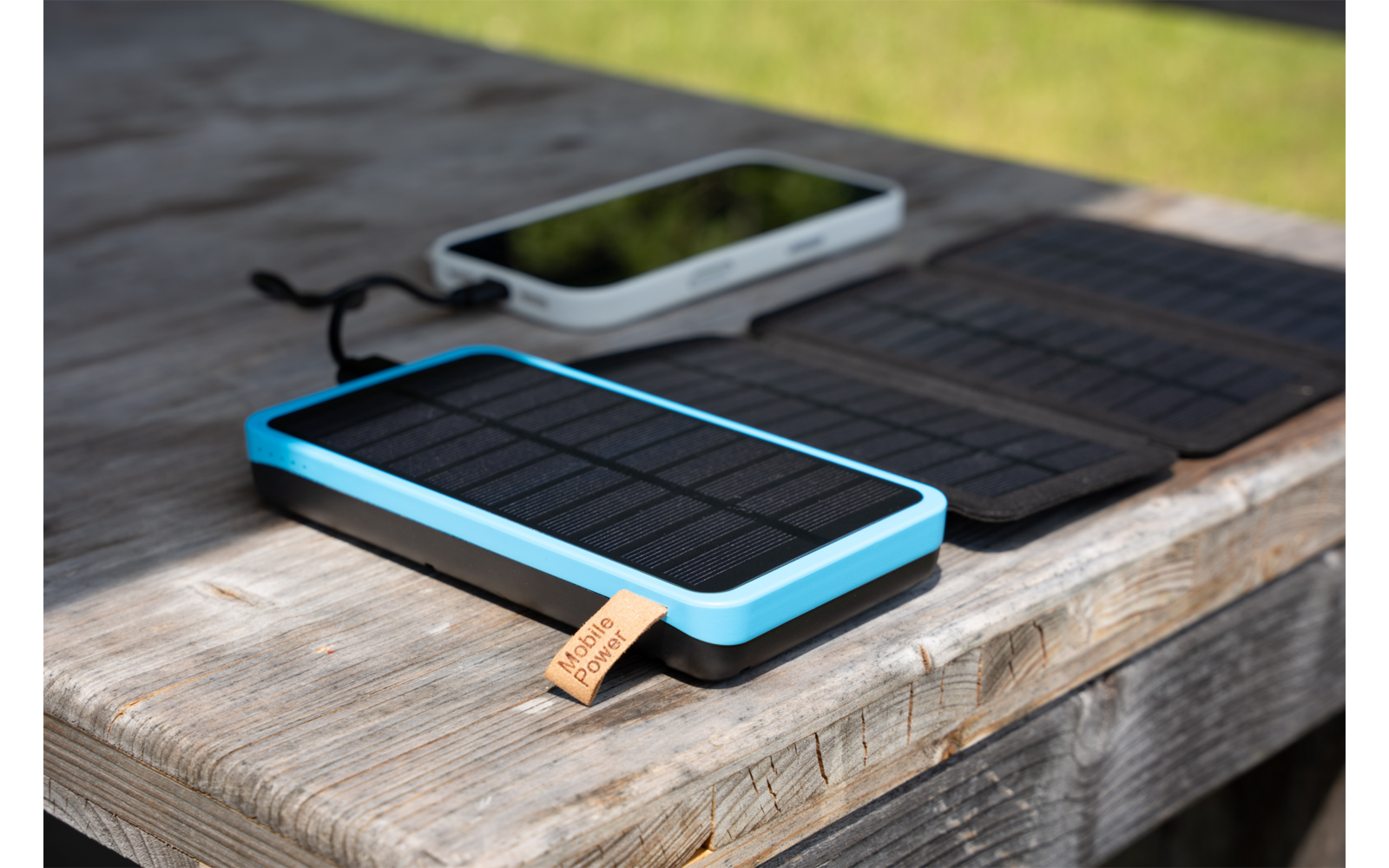 Rechteckiges Gerät mit schwarzer Solarfläche, blauem Rahmen und Lederlasche; dahinter gefaltetes schwarzes Solarpanel und weißes Smartphone mit schwarzem Bildschirm; alles auf einem verwitterten Holztisch; unscharfer grüner Rasen im Hintergrund