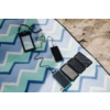 Smartphone mit Kabel, verbunden mit faltbarem Solarpanel auf blau-grünem Zickzack-Strandtuch am Sand