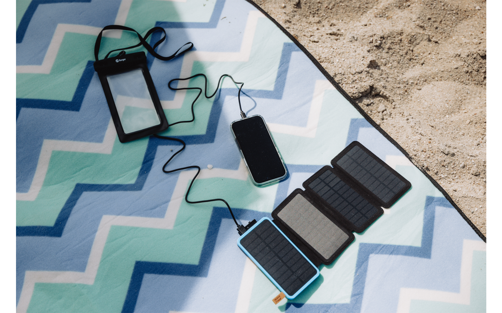 Smartphone mit Kabel, verbunden mit faltbarem Solarpanel auf blau-grünem Zickzack-Strandtuch am Sand