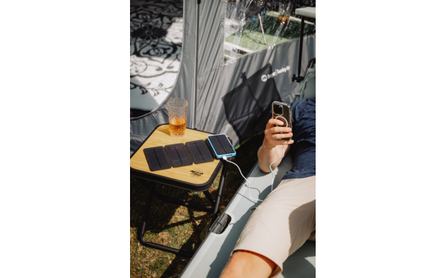 Person liegt auf aufblasbarer Liege, trägt dunkelblaues Shirt und helle Shorts, hält ein Smartphone; kleiner Holztisch neben der Liege mit zusammengefalteten schwarzen Platten und einem kleinen schwarz-blauen Panel, das über ein weißes Kabel mit dem 