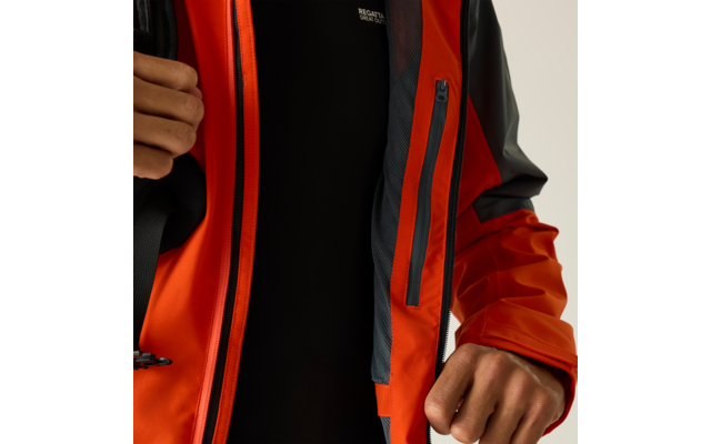 Nahaufnahme des Oberkörpers einer Person in orangeroter Regenjacke, Jacke teilweise geöffnet, darunter ein schwarzes Oberteil mit kleinem weißen Text; zwei Hände halten die Jacke und zeigen einen Reißverschluss sowie eine senkrechte Innentasche