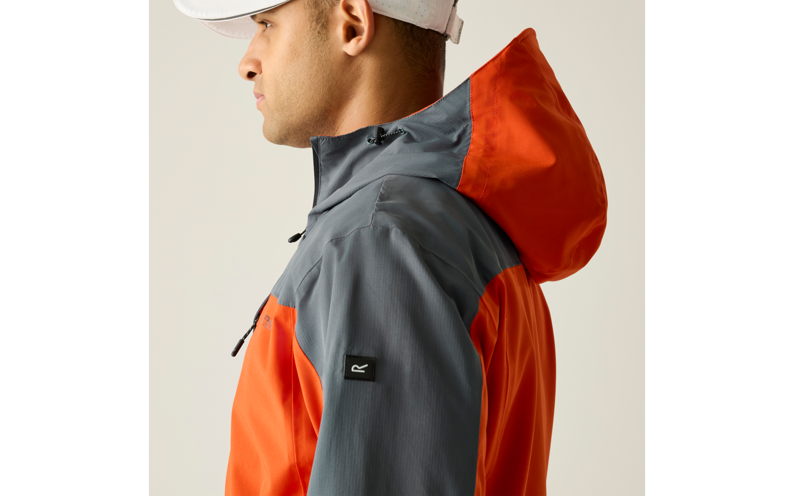 Person im Profil mit weißer Kappe und orange-grauer Kapuzenjacke, Reißverschluss und schwarzes Logo-Patch am Ärmel, heller Hintergrund