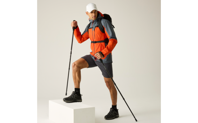 Person in orange-grauer Jacke, grauen Shorts und weißer Kappe, mit schwarzen Wanderschuhen und Rucksack, hält zwei Trekkingstöcke, linker Fuß auf weißem Quader, heller einfarbiger Hintergrund