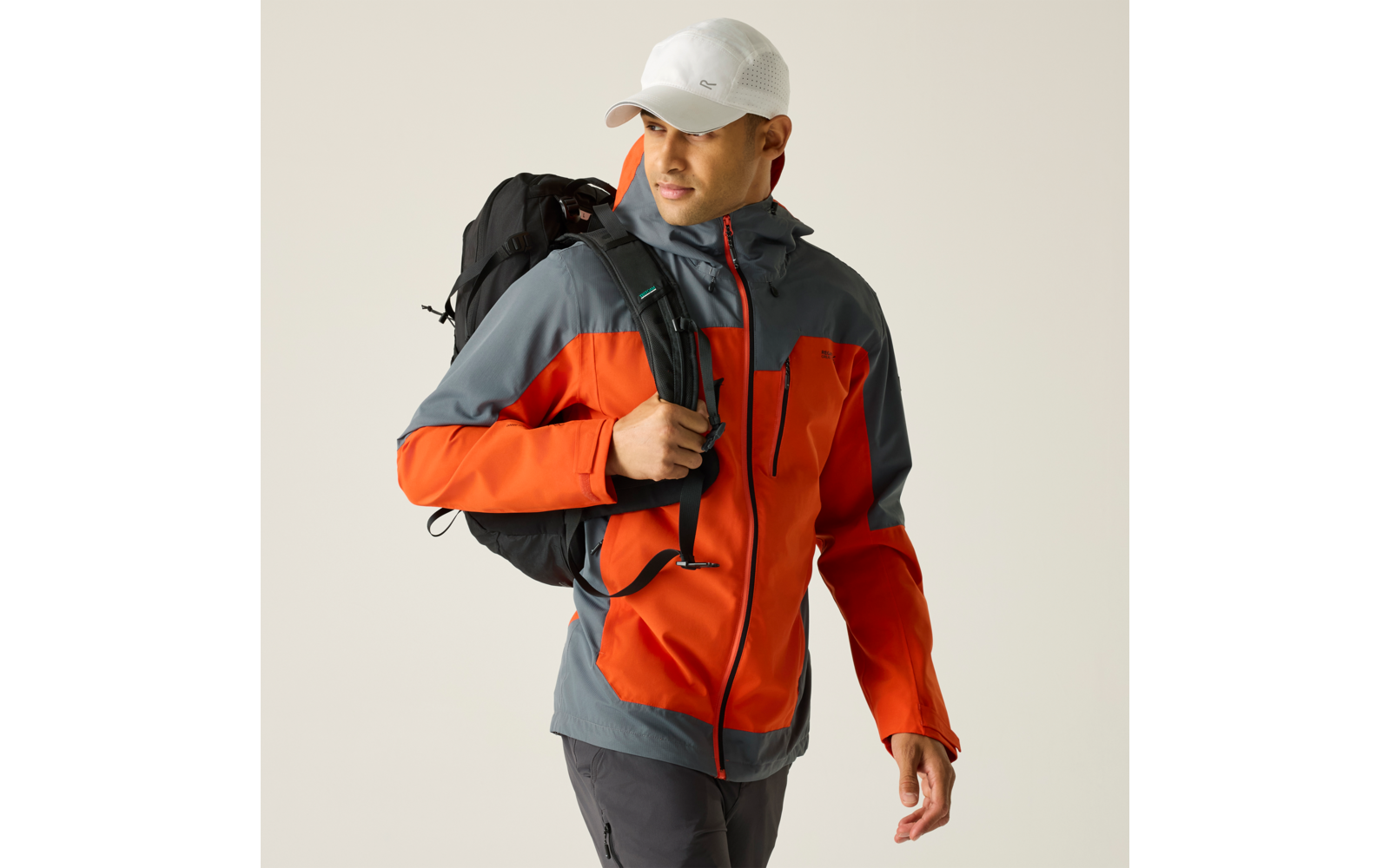 Person in orange-grauer Outdoorjacke und weißer Kappe trägt einen schwarzen Rucksack und schaut nach rechts