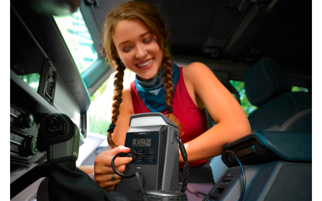 Jonge vrouw met gevlochten haar en rood topje leunt in een voertuig en steekt een kabel in een grijs apparaat met stopcontacten en display