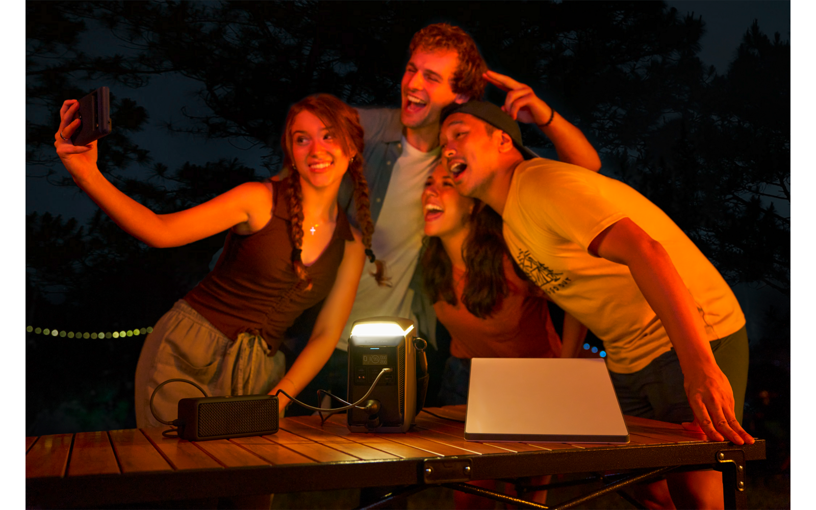 Vier personen maken een selfie; één persoon houdt een smartphone op armlengte. Op de houten tafel voor hen staan een lamp, een laptop en een luidspreker; op de achtergrond donkere bomen en een lichtsnoer