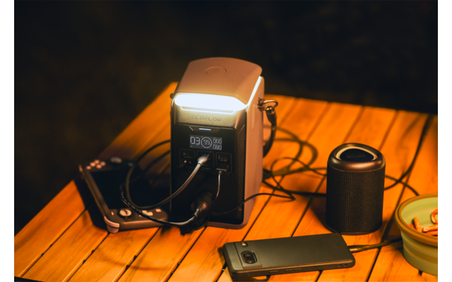 Draagbaar stroomstation met ingeschakeld licht en aangesloten kabels op een houten tafel; daarnaast smartphone, camera, cilindervormige speaker en klein bakje