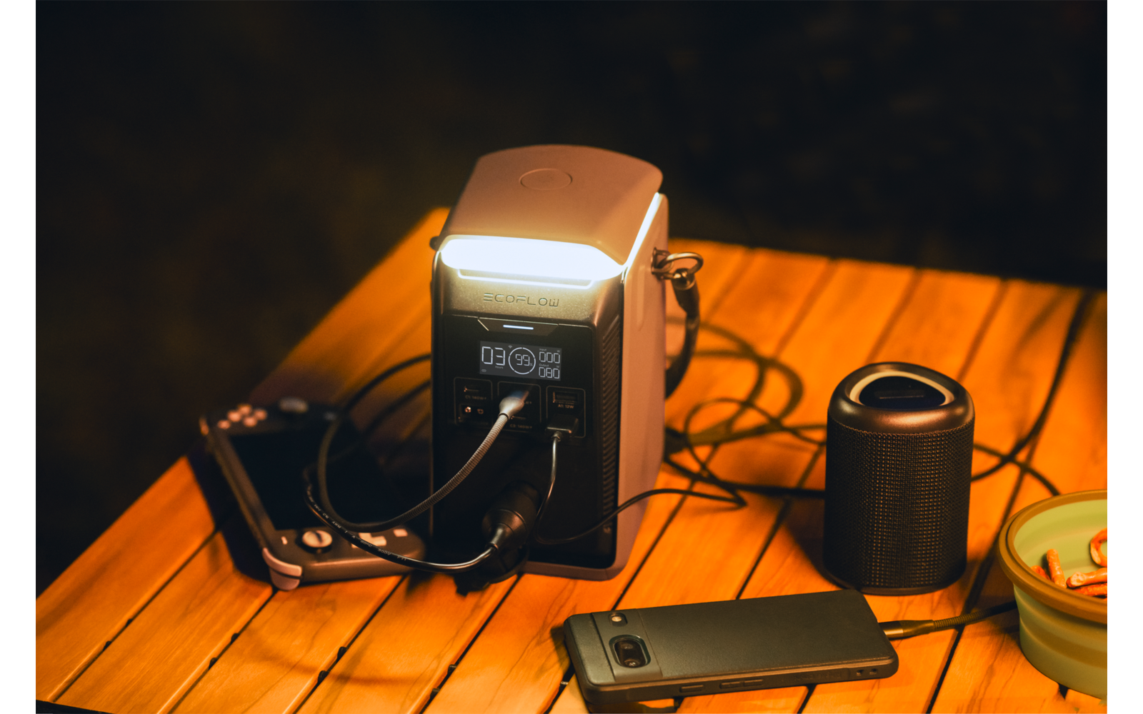Draagbaar stroomstation met ingeschakeld licht en aangesloten kabels op een houten tafel; daarnaast smartphone, camera, cilindervormige speaker en klein bakje