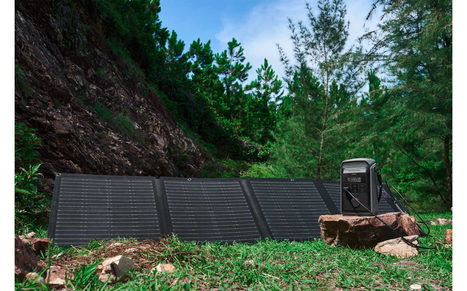 Uitgevouwen opvouwbare zonnepanelen op gras; rechts op een rots een zwarte draagbare powerstation; bomen en blauwe lucht op de achtergrond