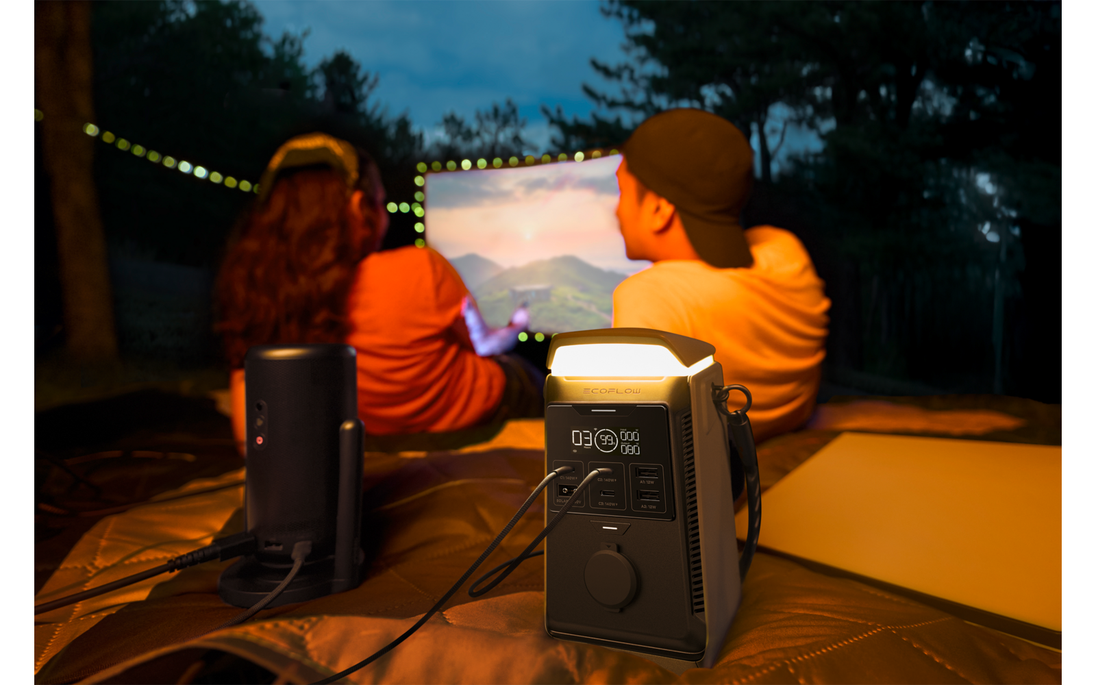 Twee personen van achteren zittend op een deken buiten voor een projectiescherm; op de voorgrond een draagbaar stroomstation en een luidspreker, lichtsnoer en bomen op de achtergrond