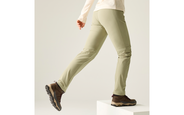 Unterkörper einer Person in hellgrünen Hosen und braunen Wanderschuhen, ein Fuß auf einem weißen Block, heller einfarbiger Hintergrund